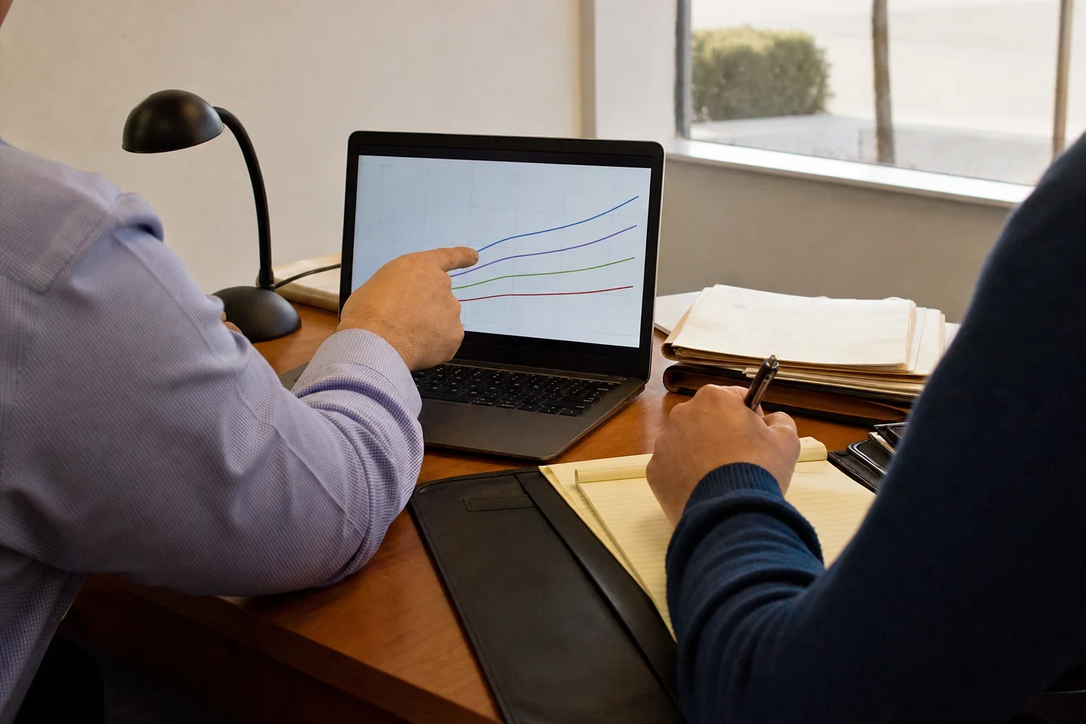 Agent in a lavender shirt points to a line chart on a laptop while a client in a navy sweater takes notes on a legal pad at an office desk with folders and a lamp