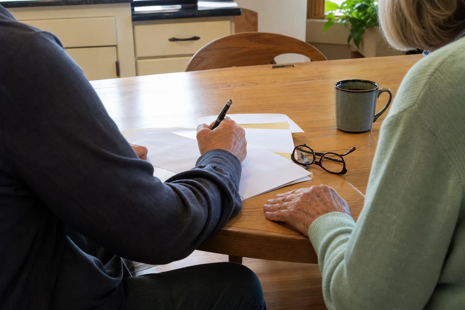 Two people seated at a wooden kitchen table, one writing on blank papers with a pen, an older woman in a mint sweater beside reading glasses and a coffee mug